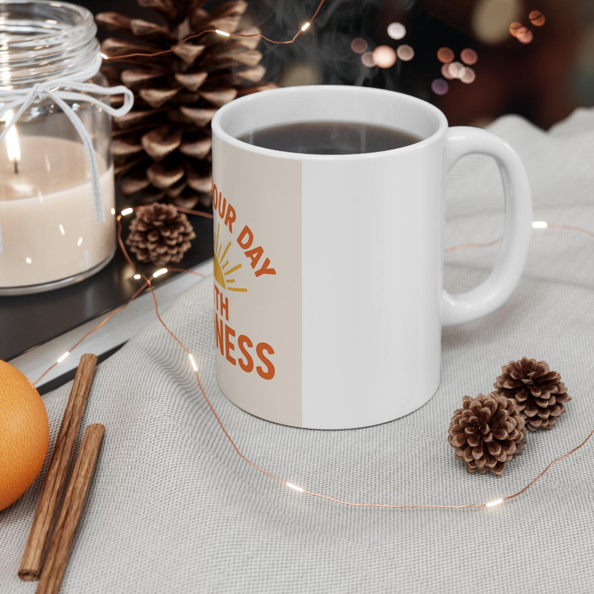 White kindness mug filled with a hot drink beside a candle, pinecones, cinnamon sticks, and string lights.